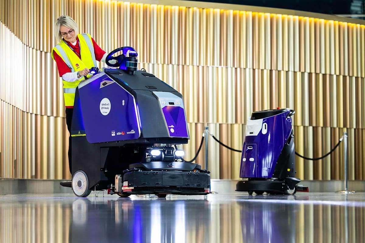 Two purple and black Mitie cleaning robots in London Heathrow Airport, with a female Mitie worker in a high vis vest using the right-hand robot